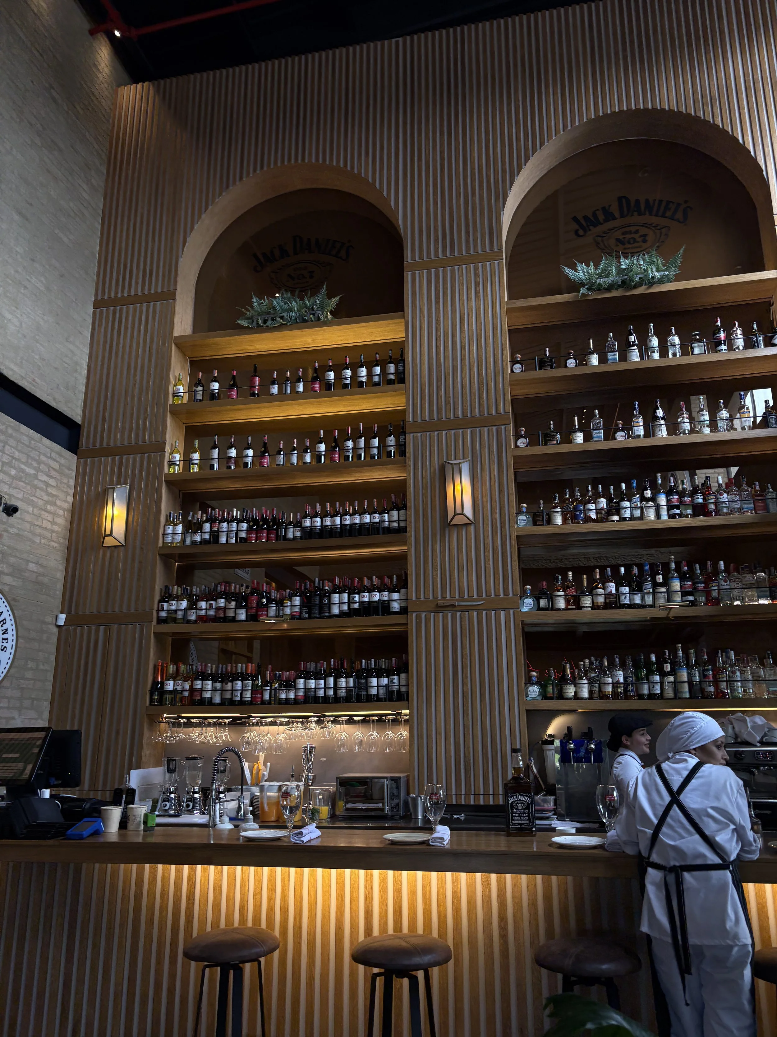 A bar with a tall wooden shelf holding numerous bottles of alcohol, two arched niches with Jack Daniel's branding, and two staff members preparing drinks behind the counter.