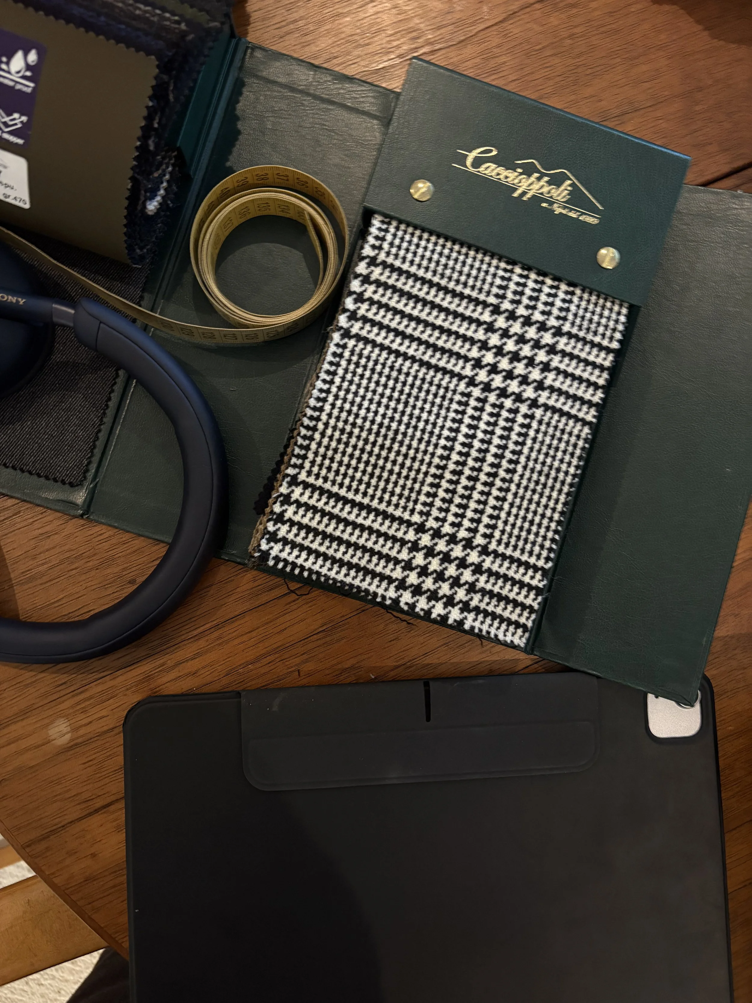 Fashion swatch book with black and white houndstooth fabric, corner of a laptop, yellow measuring tape, a pair of headphones, and fabric swatches on a wooden table.