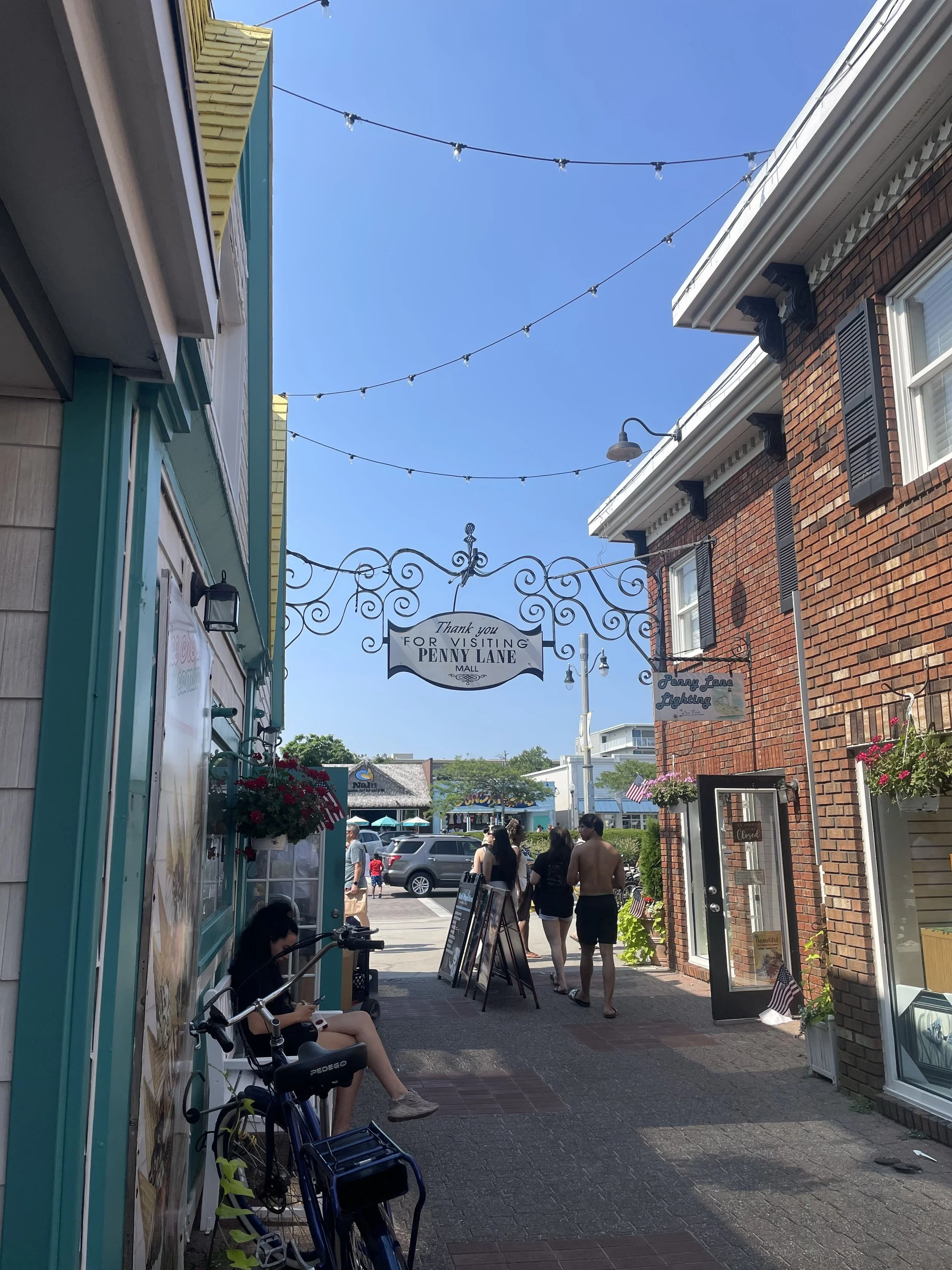 People walking down a small commercial street with colorful storefronts, hanging flower baskets, and string lights overhead, leading toward a parking lot and distant shops.