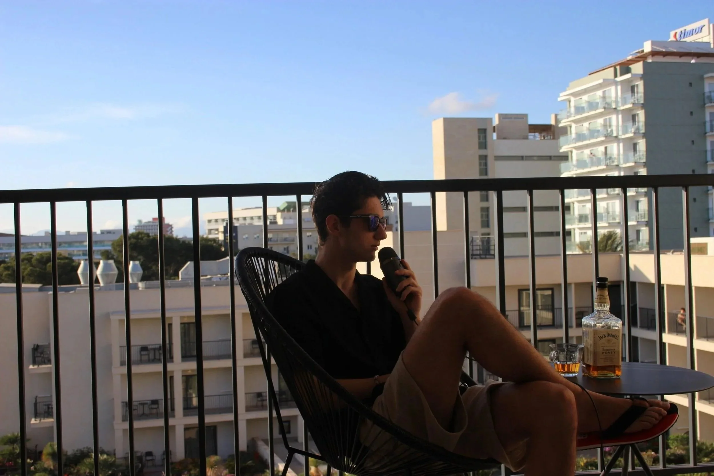 A young man with sunglasses sitting on a balcony holding a microphone, with a bottle of Jack Daniel's whiskey on a table beside him, overlooking a cityscape with modern buildings.