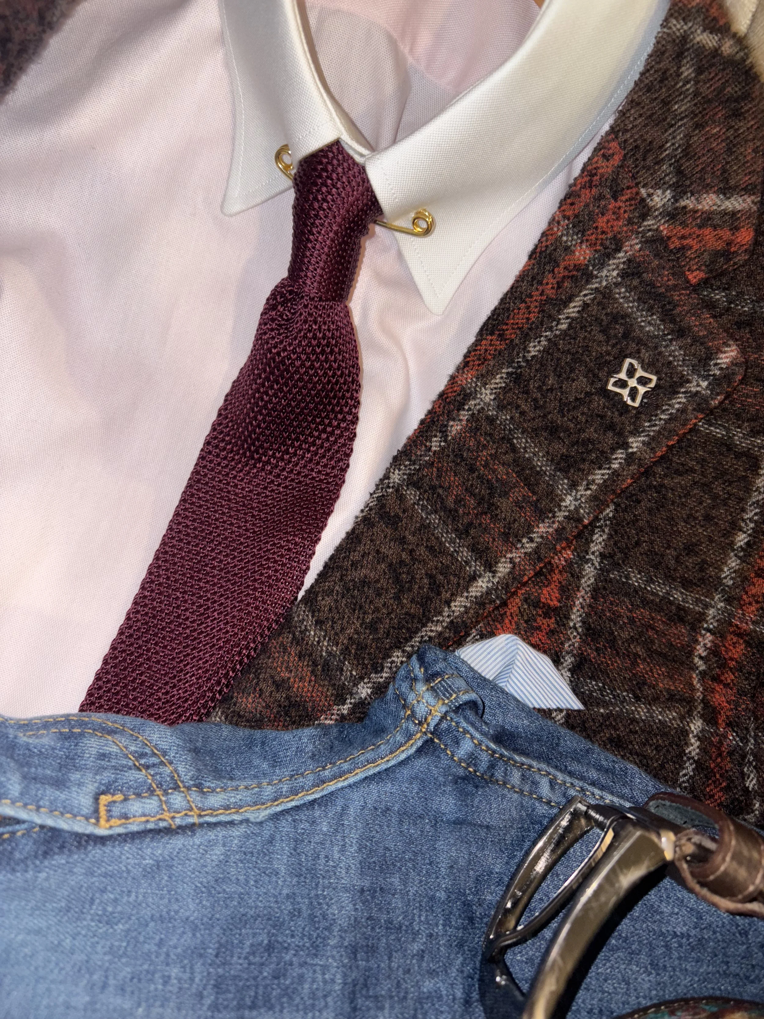 Close-up of a formal outfit including a white shirt with a burgundy tie, a plaid blazer with a lapel pin, and blue jeans with a belt.