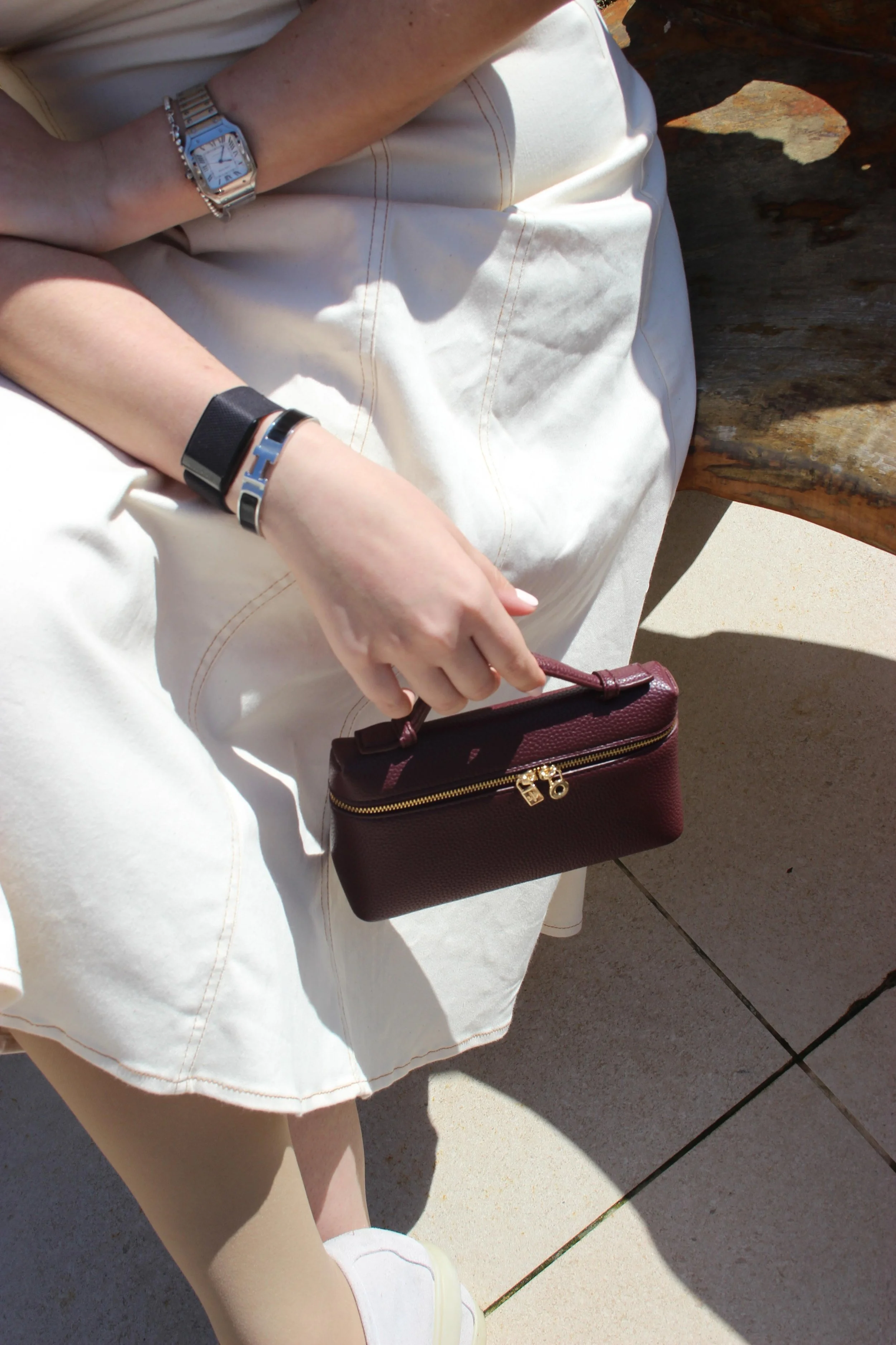 A woman dressed in white sitting on a wooden bench, holding a small dark red handbag with a gold zipper. She is wearing a silver wristwatch, a black bracelet, and a silver bracelet on her left wrist. She is also wearing beige pants and white shoes, w