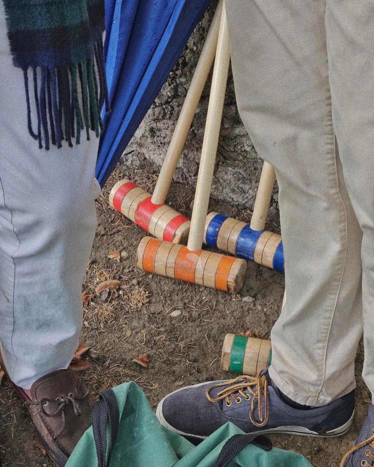 Close-up of the lower half of two people standing on dirt, with some colorful wooden toy nails and a wooden hammer on the ground between them, and a green bag at the bottom of the image.