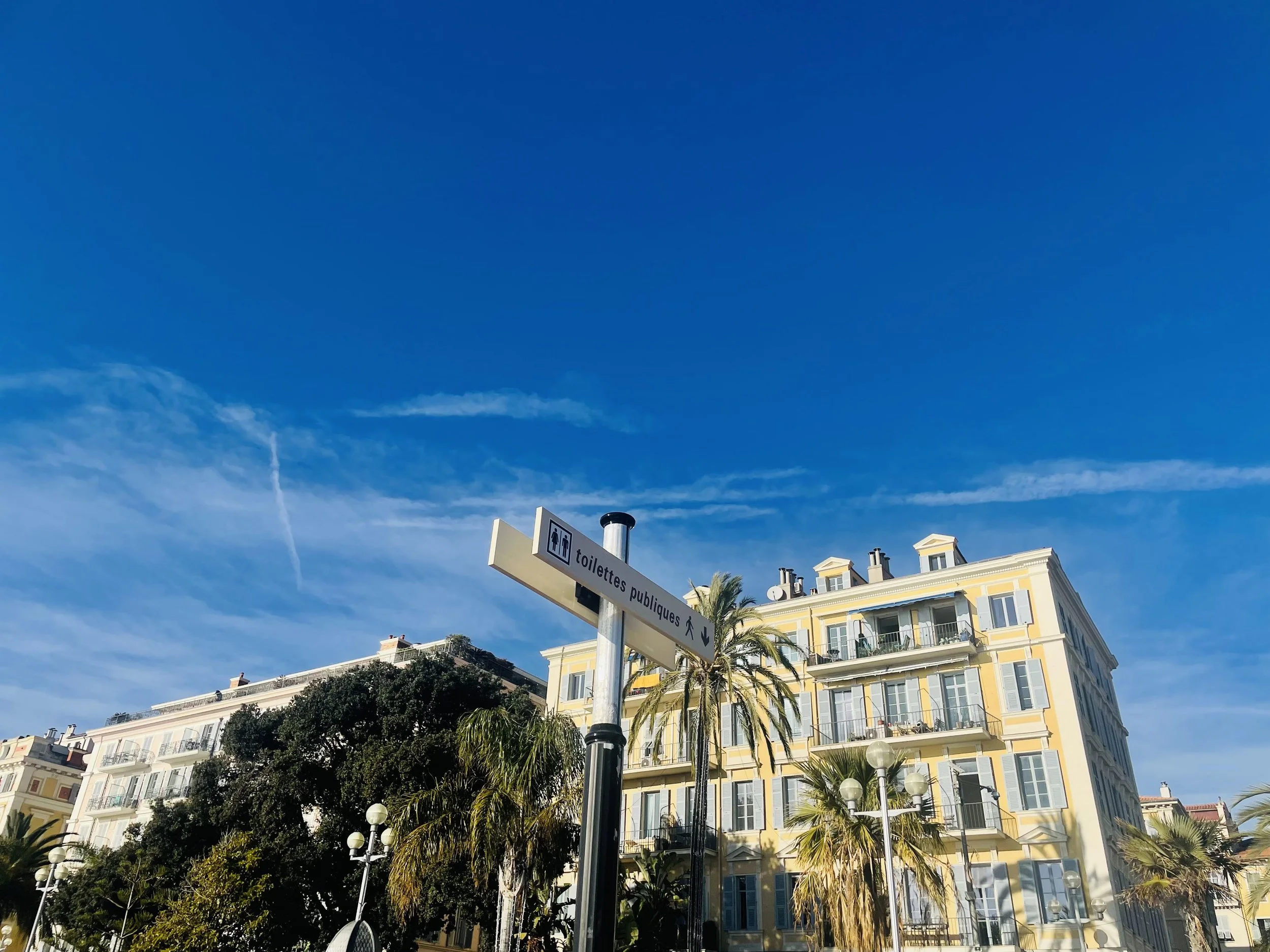 A yellow building with balconies and large windows, surrounded by palm trees, under a clear blue sky with some clouds, and a sign indicating public restrooms.