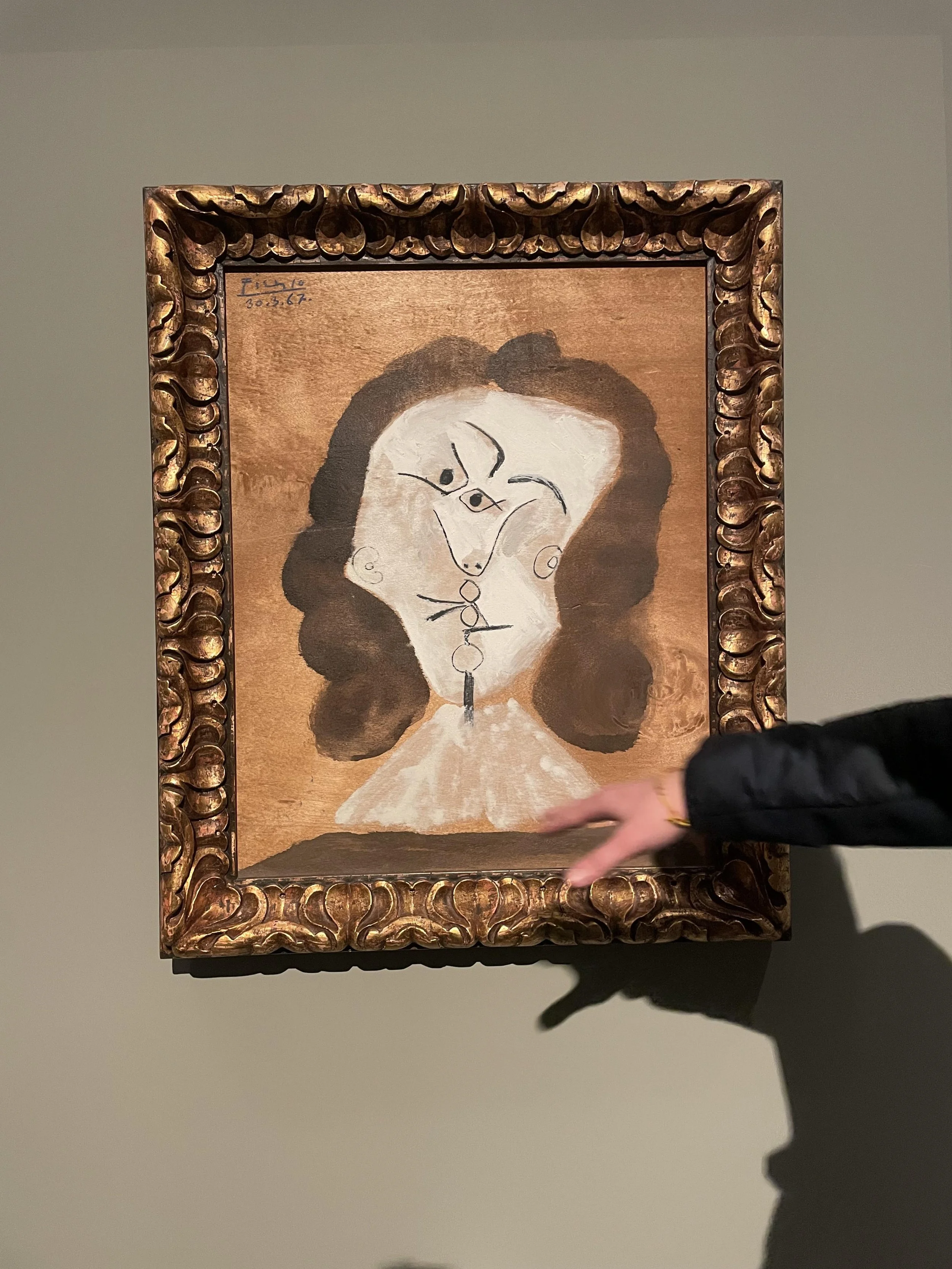 A framed painting of a woman with dark curly hair, painted in an abstract and minimalist style, hanging on a plain wall. A person’s hand is pointing towards it.
