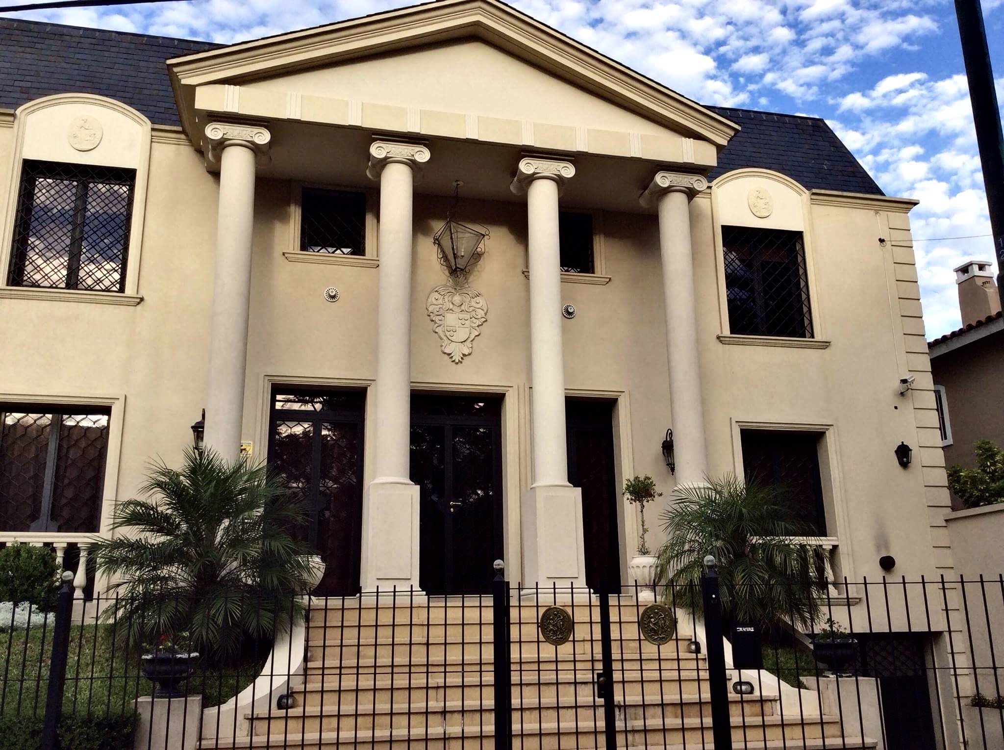 Front exterior of a grand, cream-colored two-story house with classical architectural elements including columns, a pediment, and decorative carvings, with stairs leading up to the entrance and a black fence in front.