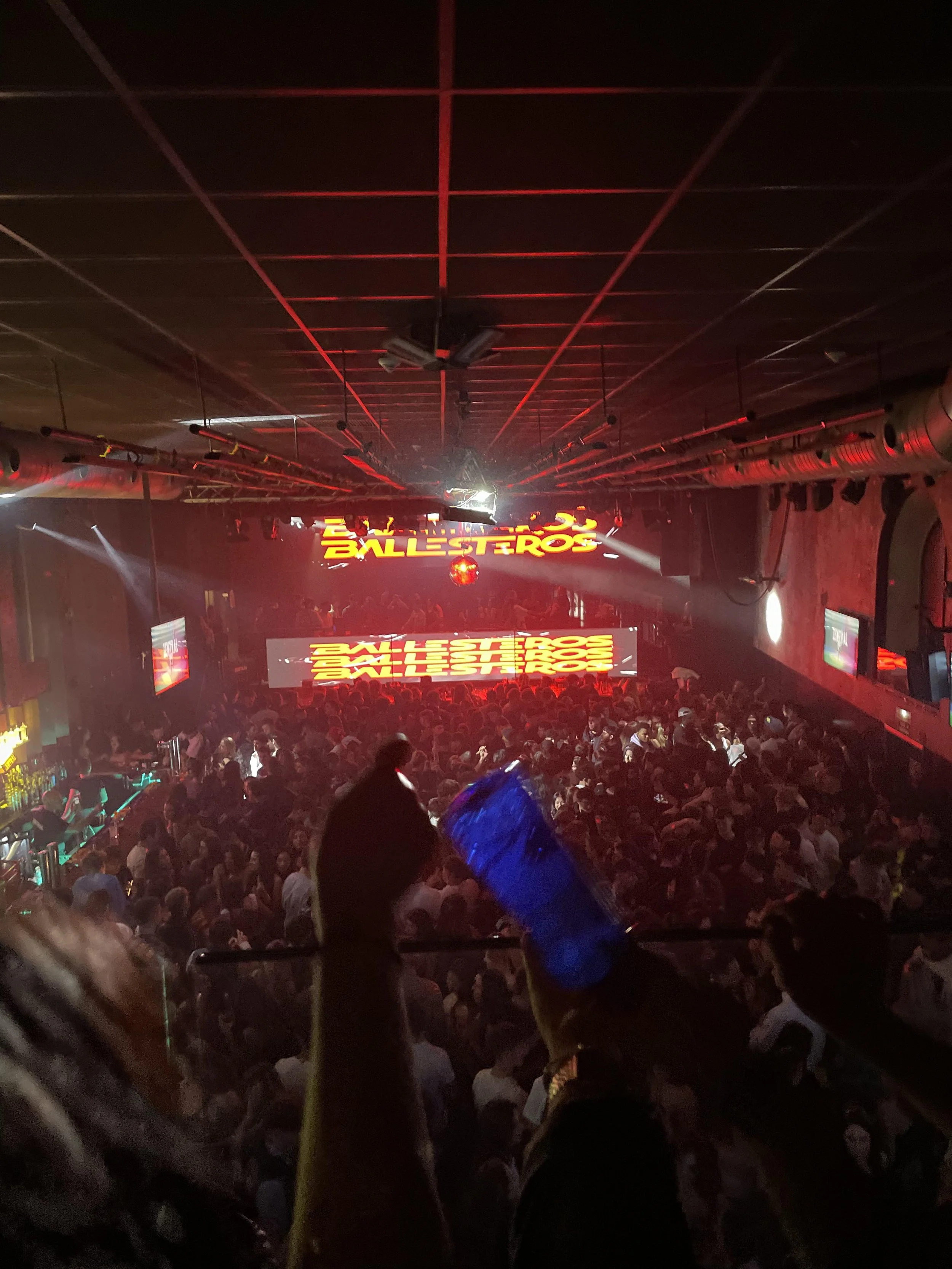 Crowded nightclub with a stage, neon signs, and dancers, with person holding a blue drink in the foreground.