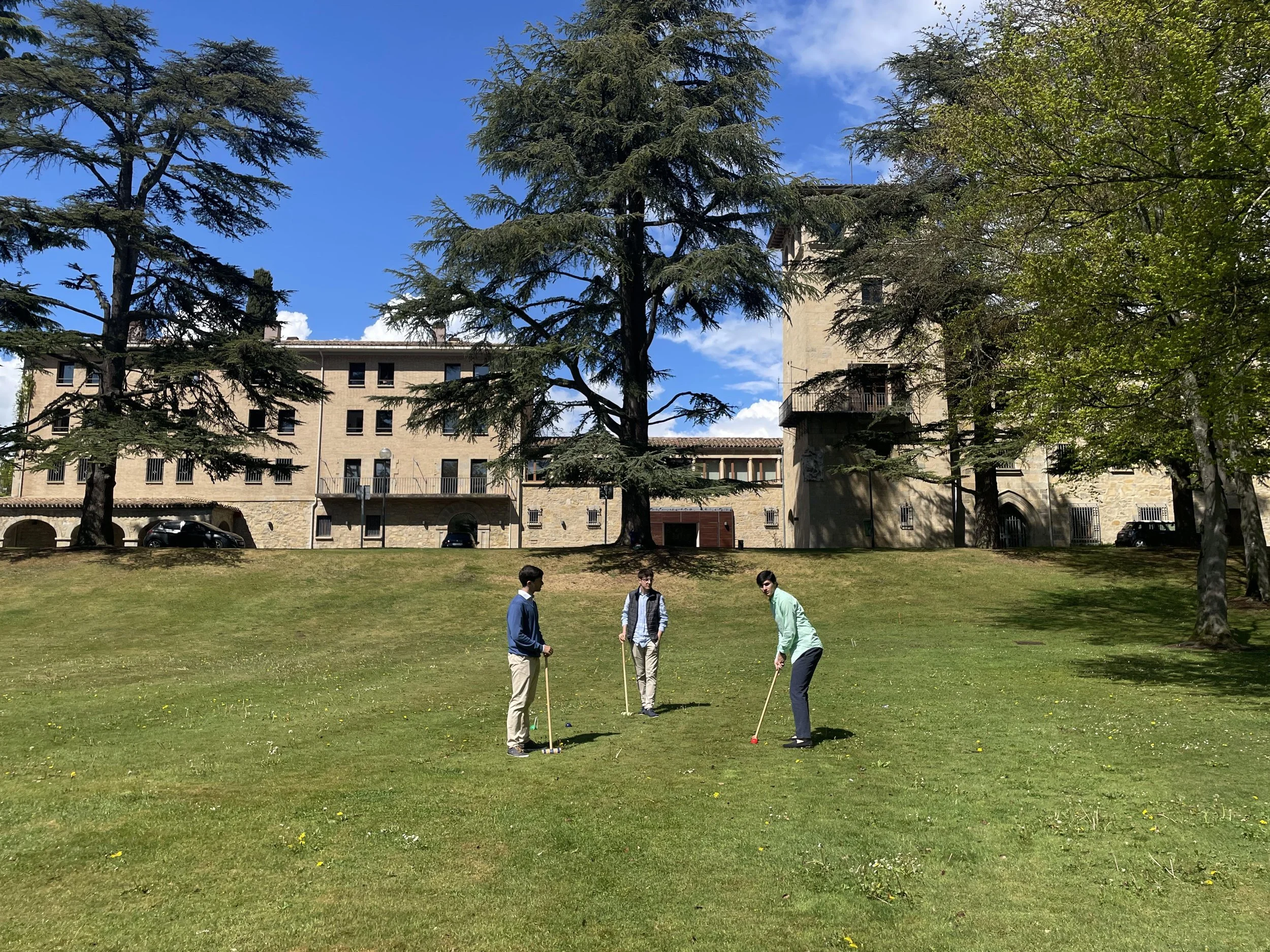 Three young men playing mini golf on a grassy field with trees and a large stone building in the background under a partly cloudy sky.