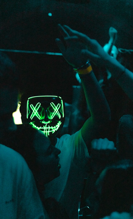 Person wearing a neon skull mask at a dark party or club with their arms raised.
