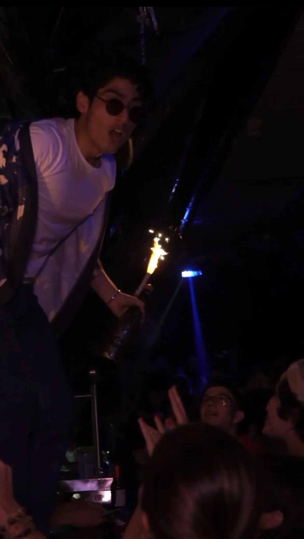 A young man with sunglasses and a backpack holding a sparkler at a dark party or celebration with other people clapping and looking at him.