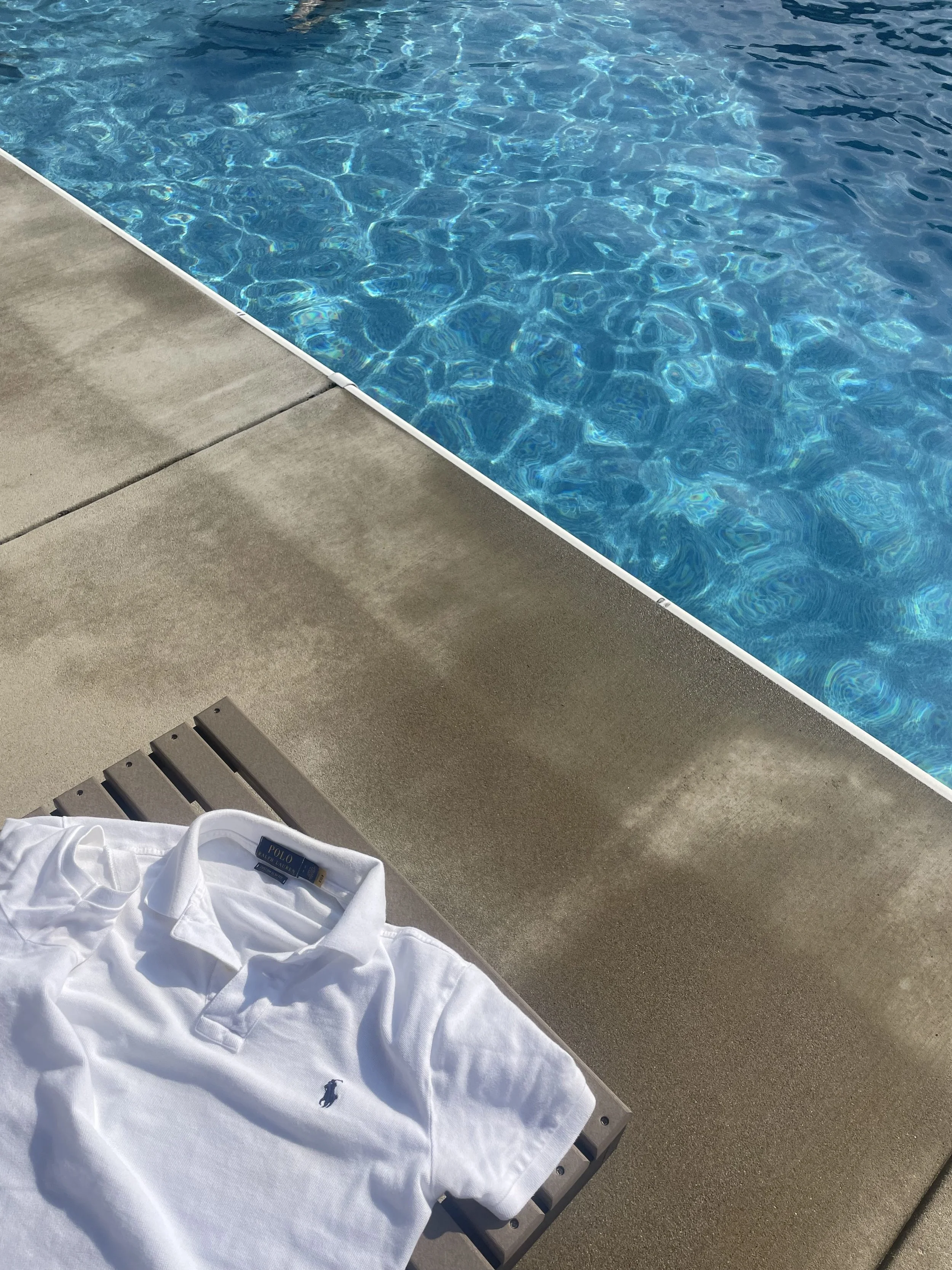 A white polo shirt with a small logo on the chest lying on a wooden lounge chair next to a swimming pool with clear blue water.
