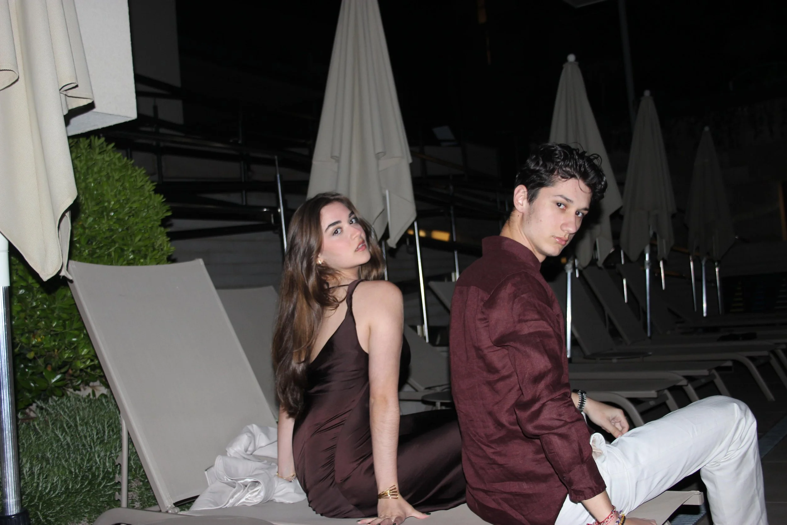 A young man and woman sitting on lounge chairs outdoors at night, with umbrellas and greenery around.