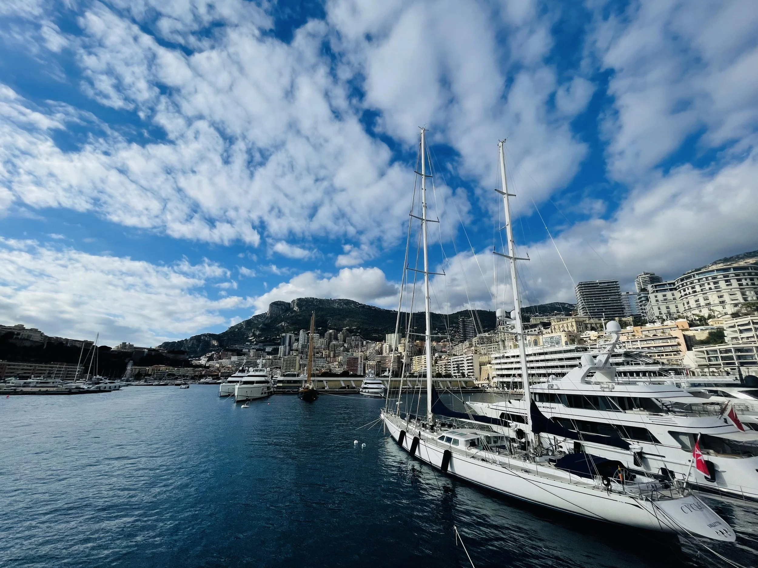 A harbor with yachts docked and a cityscape with high-rise buildings and mountains in the background under partly cloudy sky.
