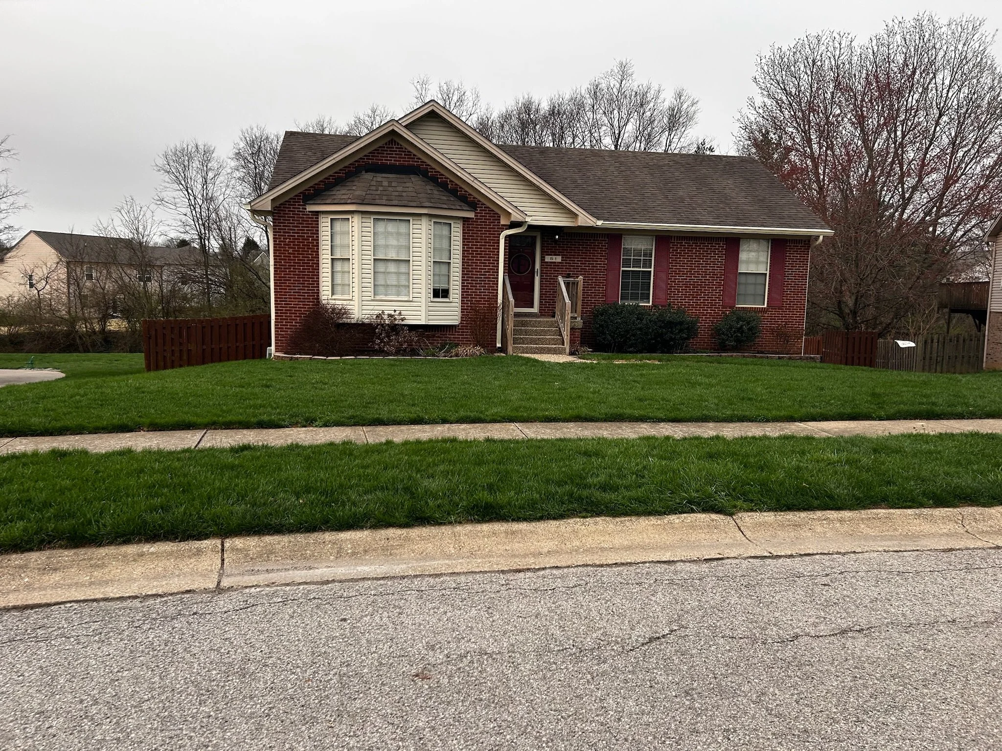 Front view of a brick house with a gabled roof, a small set of stairs leading to the front door, and a lawn with a sidewalk in the foreground. Trees without leaves are in the background, indicating late fall or winter.