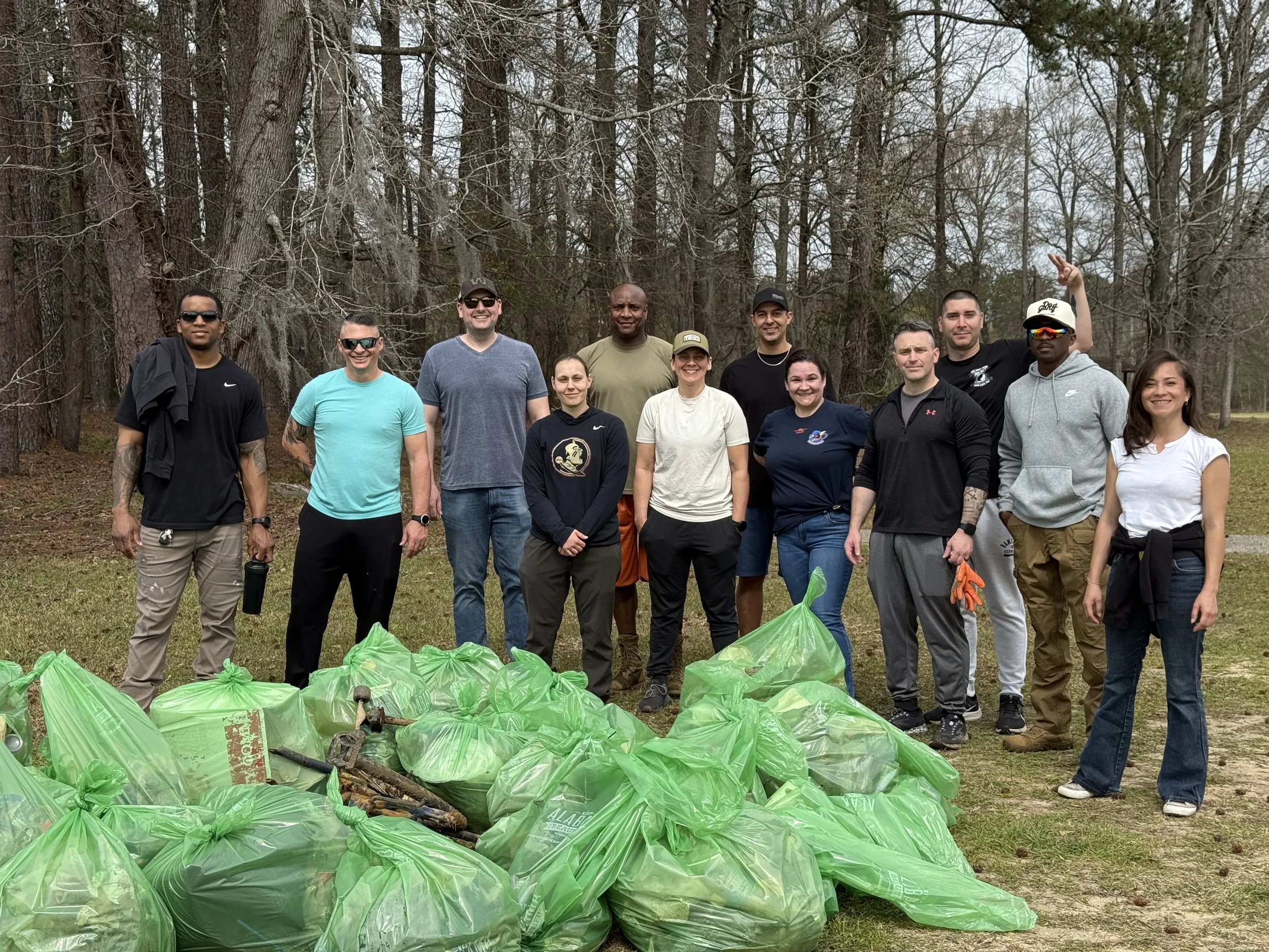 Group of 14 people standing outdoors near a pile of collected trash in green trash bags, in a wooded area with trees in the background. At Wares Ferry Park