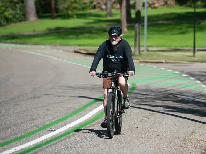 An older man with a beard and glasses riding a mountain bike on a paved park path, wearing a black helmet, black long-sleeve shirt, and shorts, with green trees and grass in the background. On the Cloverdale Trail in Montgomery, AL