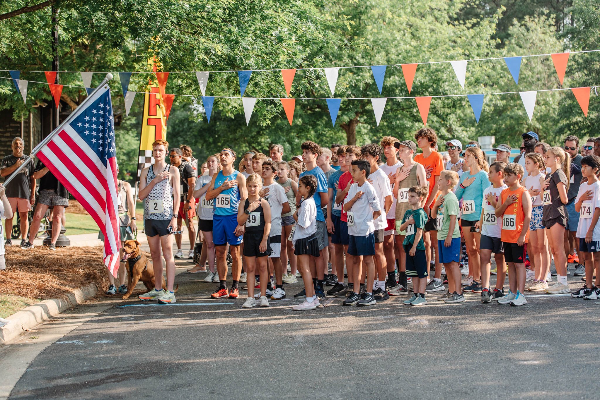 Children and adults participating in a patriotic event outdoors, standing with hands over hearts, with American flags and colorful bunting overhead for the Woodland Creek Memorial Day 5k
