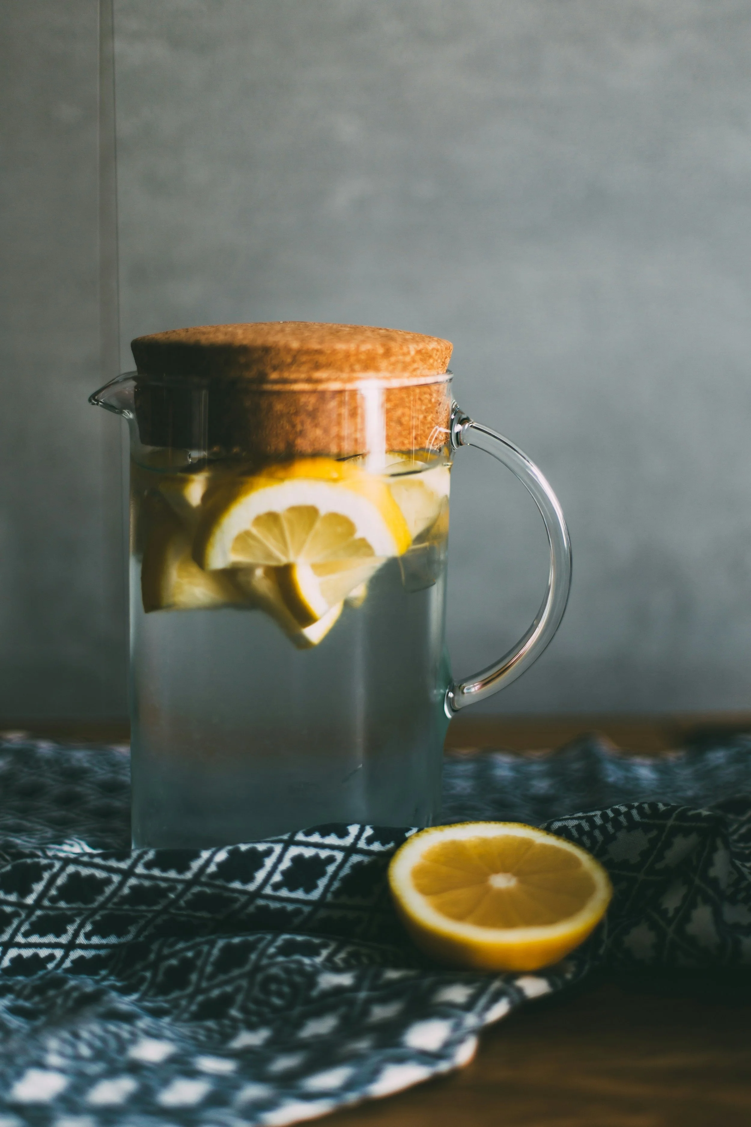 Glaskanne mit Zitronenwasser und Korkdeckel, umgeben von einem schwarzen und weißen Tuch, halbierte Zitrone im Vordergrund.