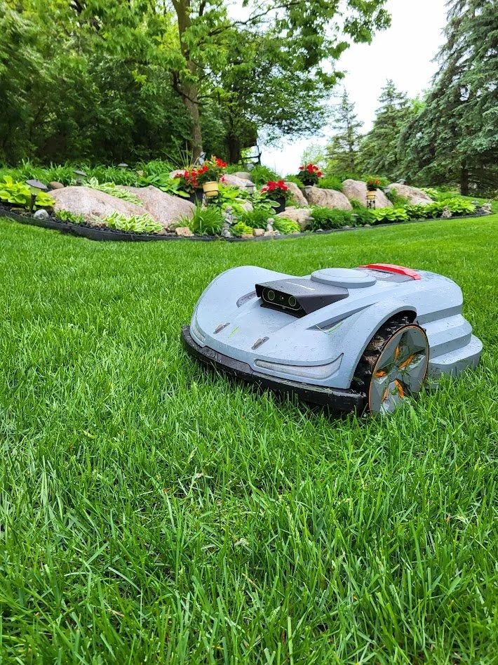 An autonomous lawn-mowing robot on a grassy yard with a garden and trees in the background.