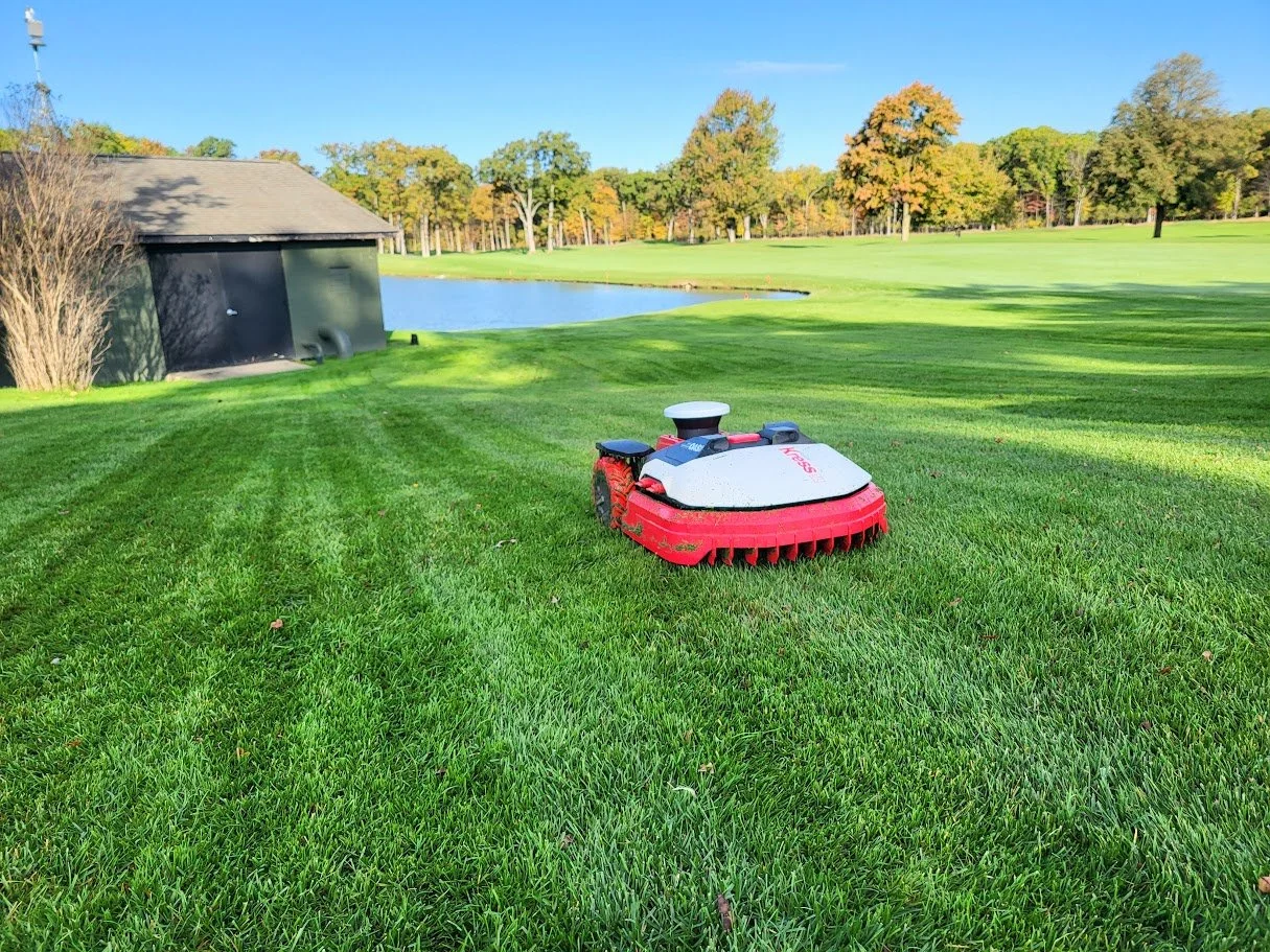 Lawn mower on a well-manicured grass field near a pond, with trees and a building in the background, on a sunny day.