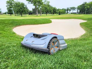 Autonomous robotic lawn mower on a grassy golf course surrounded by trees and sand trap.