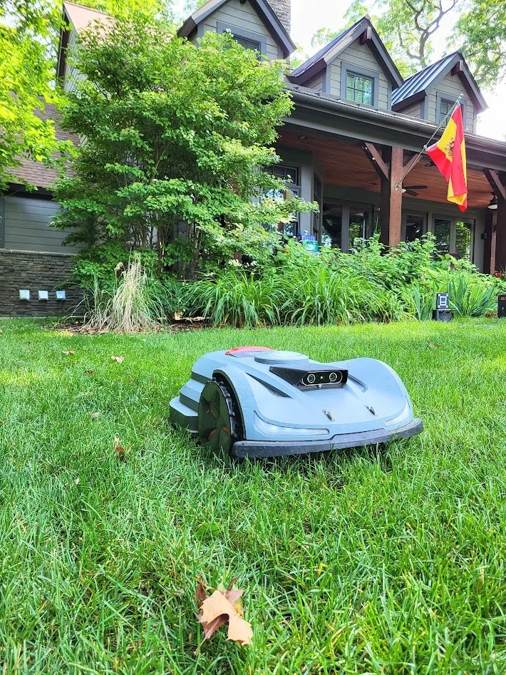 A robotic lawn mower is on the grass in front of a house with a porch, trees, and a flag, during daytime.