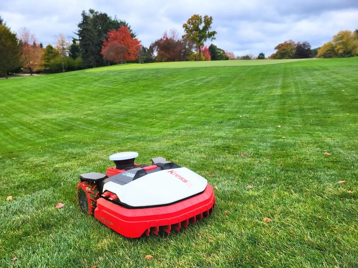 Robotic lawn mower on a well-maintained grassy field with colorful autumn trees in the background.