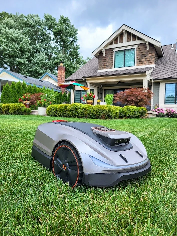 A robotic lawn mower on a well-kept lawn in front of a house with a garden, trees, and a blue sky.