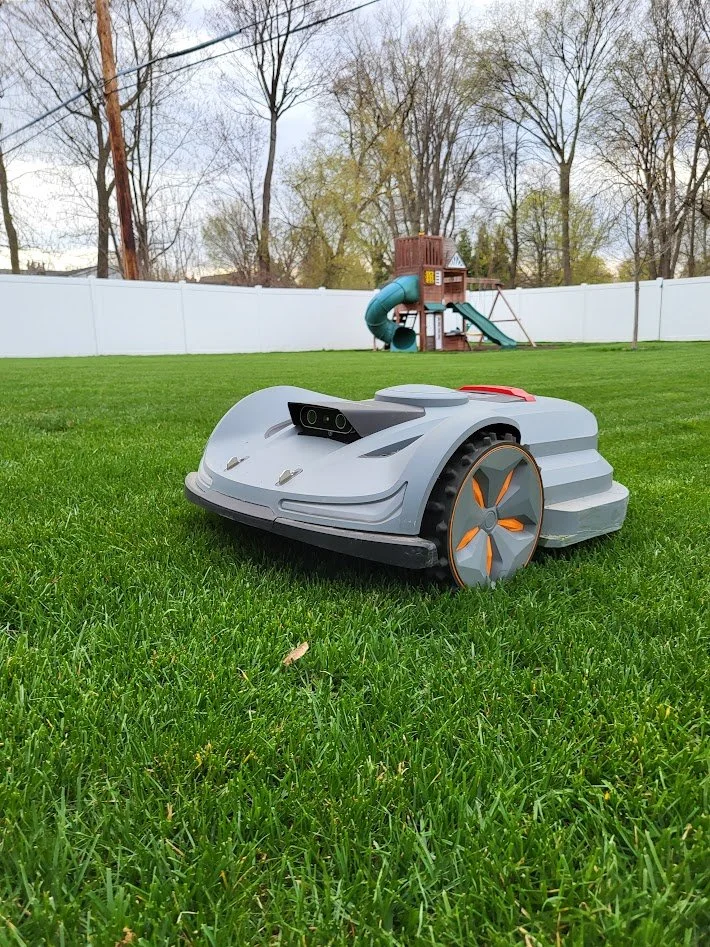 A robotic lawn mower on green grass with a backyard playground and a white fence in the background.