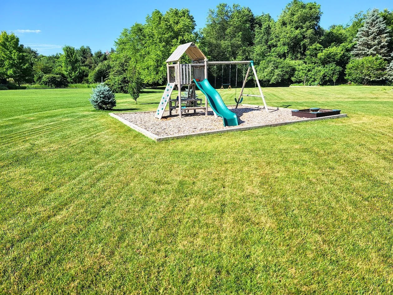 A children's playground with a slide surrounded by well-maintained green grass and trees in the background.