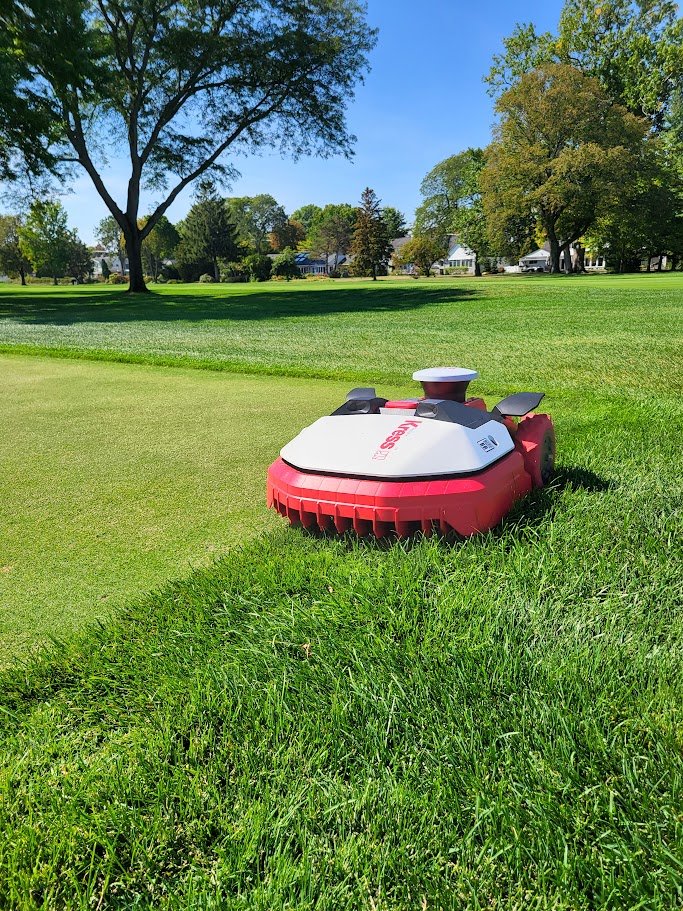 A robotic lawn mower on a well-maintained grassy area with trees and houses in the background under a clear blue sky.
