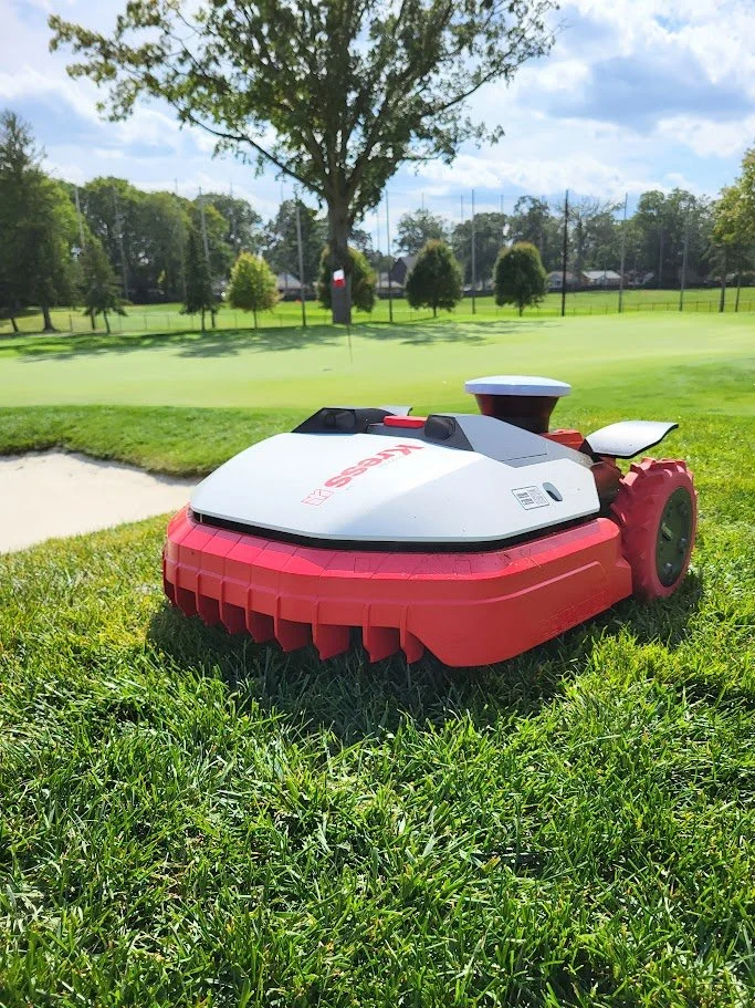A red and white robotic lawn mower on a grassy field with a park and trees in the background.