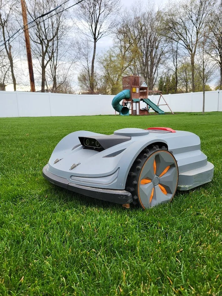 Robot lawn mower on green grass with a children's playset and trees in the background.