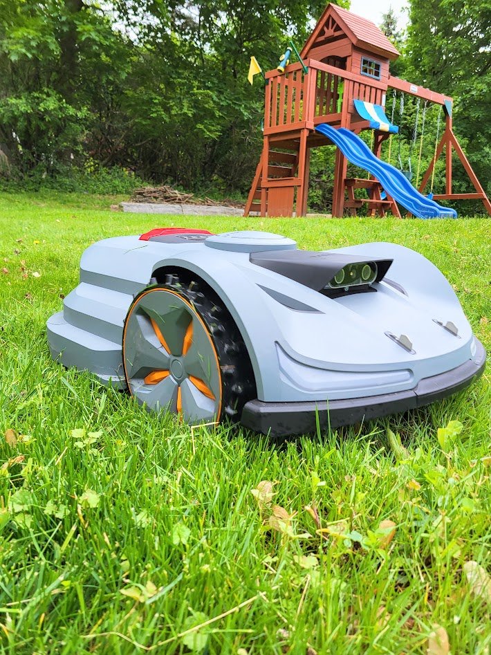 A robotic lawn mower on grass, with a wooden playset in the background.