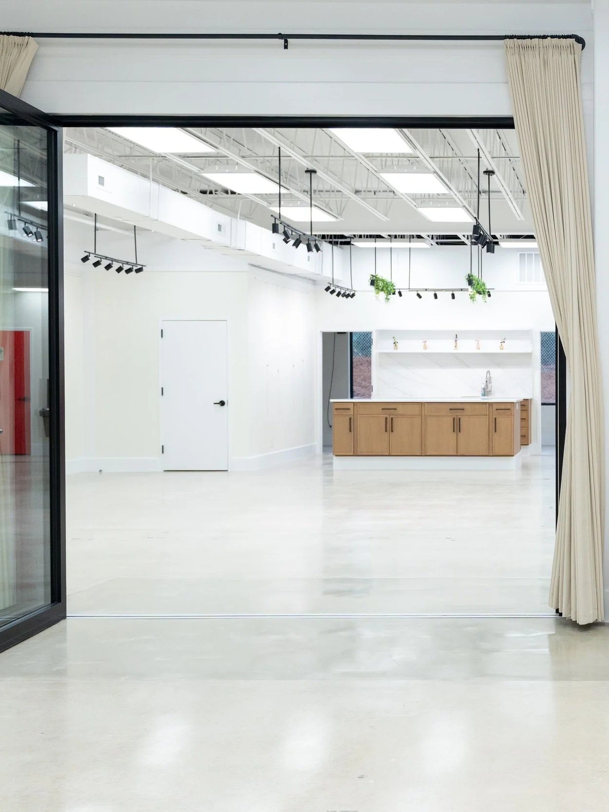 Empty art gallery space with white walls, wooden accents, colorful paintings, brown and gray chairs, red door, and large open door with curtains.