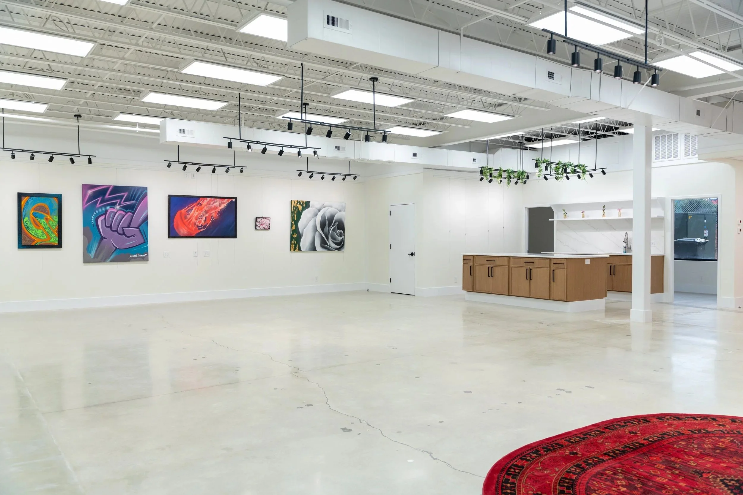 An art gallery with paintings hung on white walls, a wooden counter to the right, and a corner with plants hanging from the ceiling, all inside a well-lit space with a red rug on the floor.