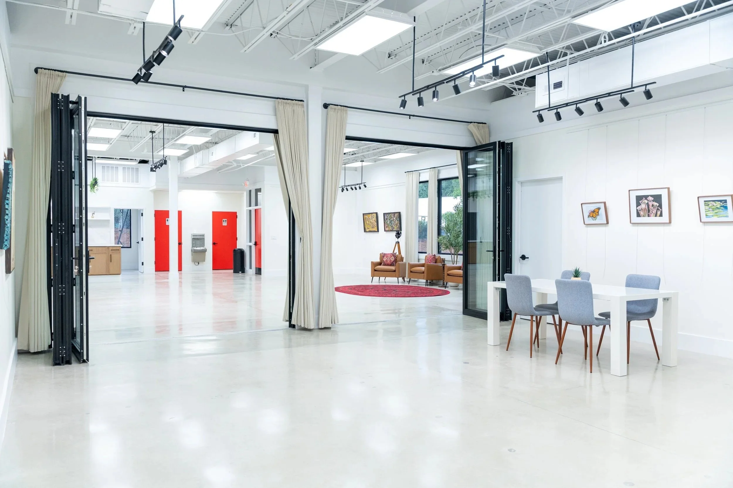 Modern, spacious interior with white walls and light flooring, featuring a sitting area with brown chairs and colorful artwork, a white dining table with gray chairs, large folding glass doors, and a bright, open background with red doors and a water