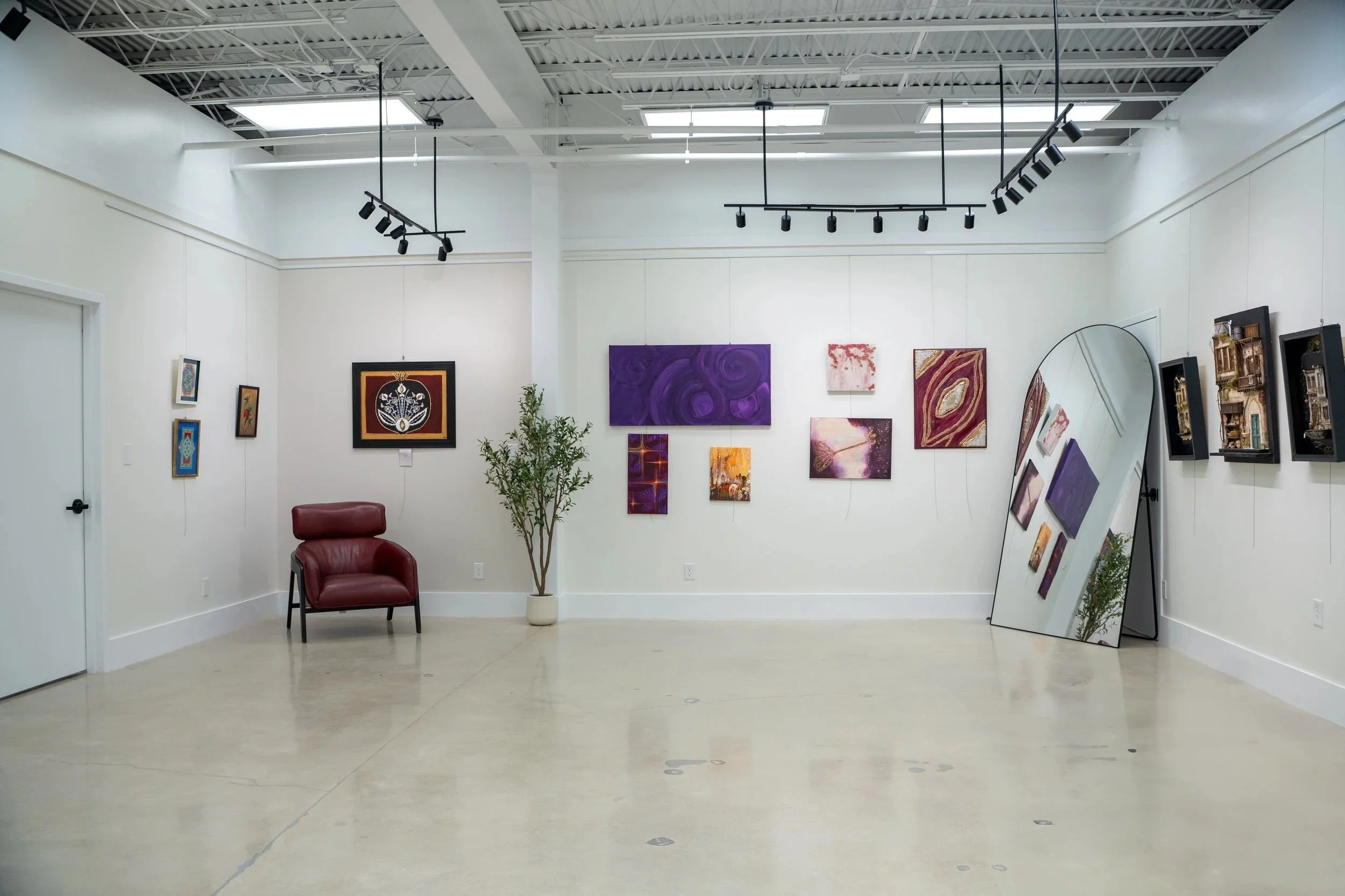 An art gallery with white walls, featuring various colorful paintings, a large mirror leaning against the wall, a maroon chair, and a potted plant. Overhead track lighting illuminates the artwork.