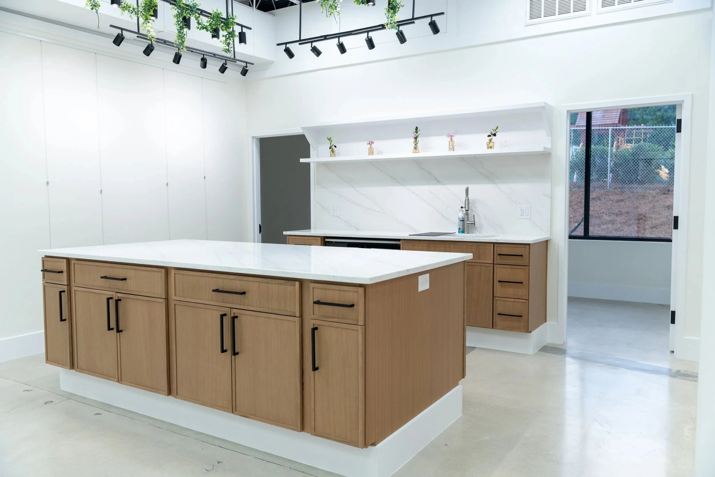 Modern kitchen with wooden cabinets, white marble countertops, a sink, black handle hardware, and a large window showing outdoor fencing and trees.