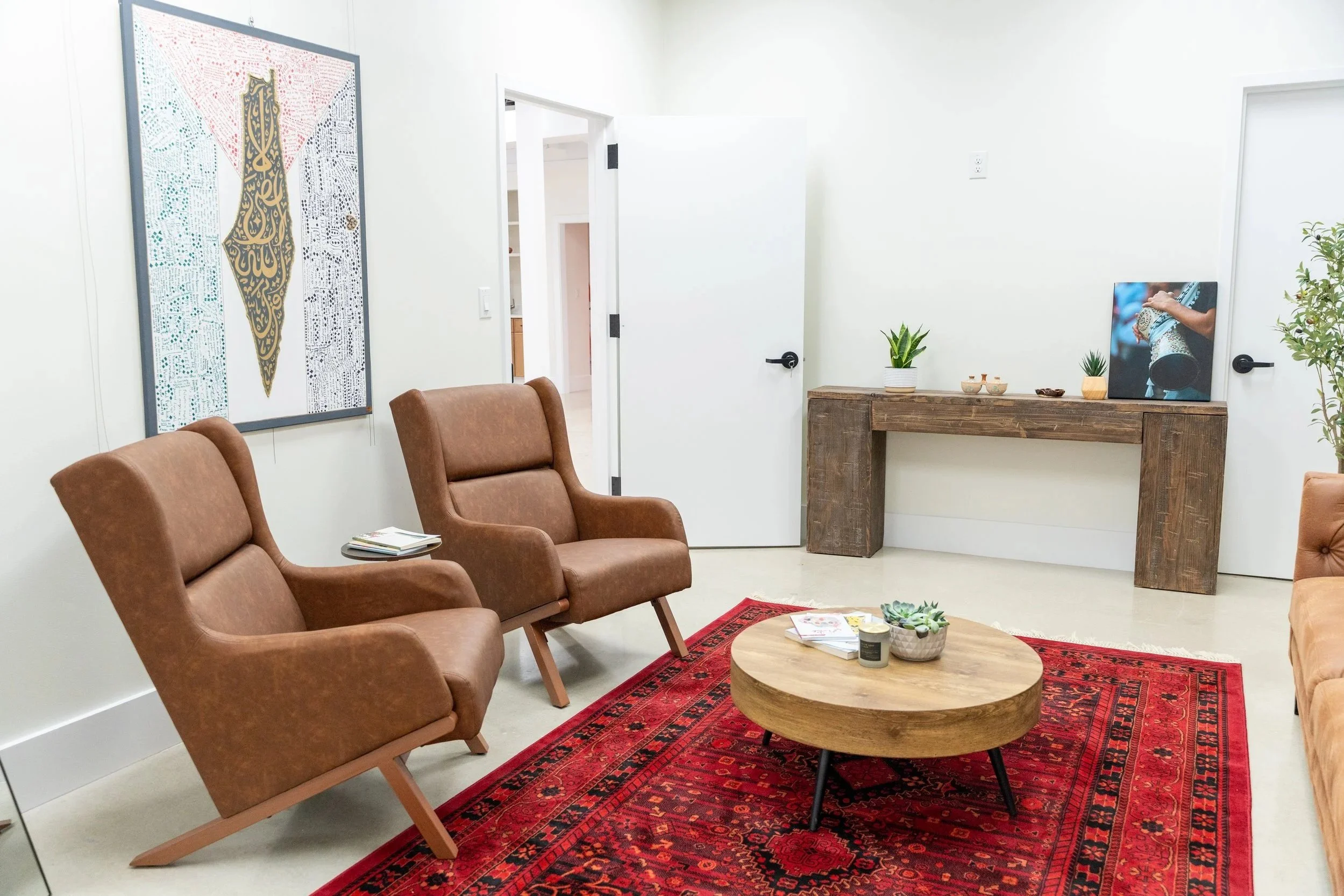 Living room with three brown leather chairs, a wooden coffee table with magazines and candles, a red patterned rug, a wooden sideboard with plants and artwork, and a white wall with a large framed art piece.