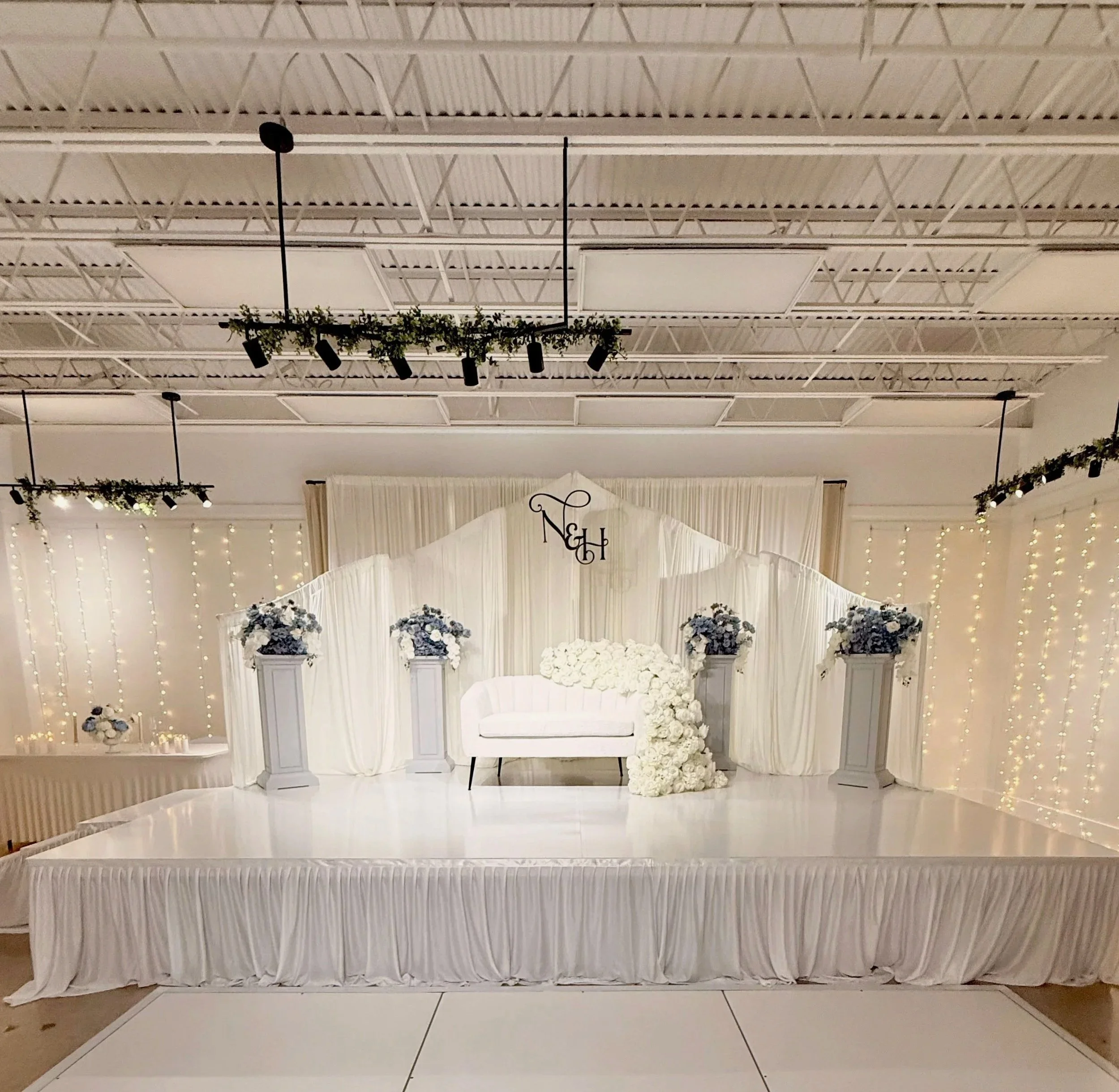 An elegant white wedding altar decorated with white flowers and greenery, with draped white fabric for the backdrop, and floral arrangements on pedestals, set up for a wedding ceremony.