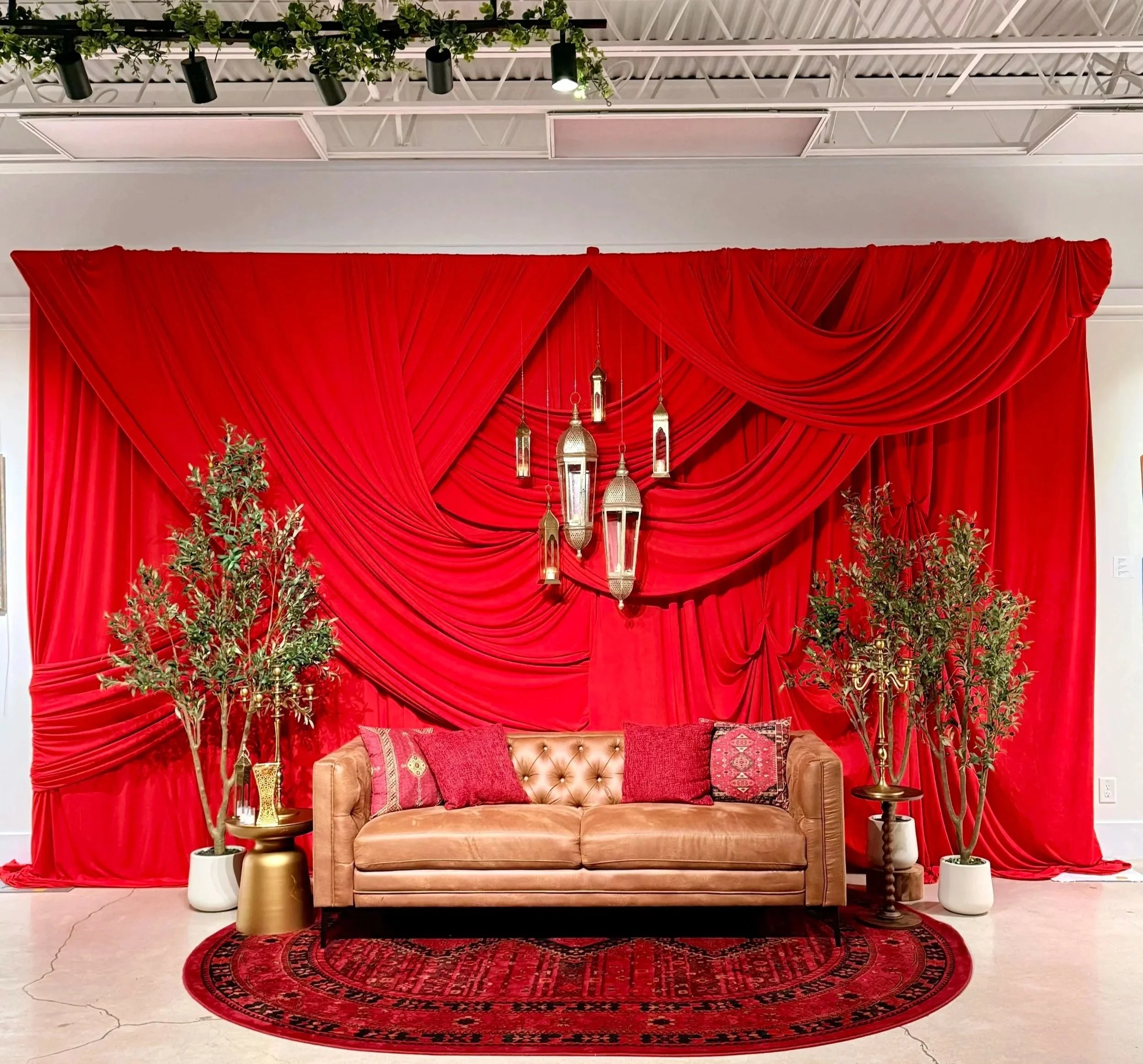 A decorated indoor space with a red draped backdrop, hanging lanterns, leafy plants, a tan sofa with red pillows, a round red rug, and side tables with vases and decorative items.
