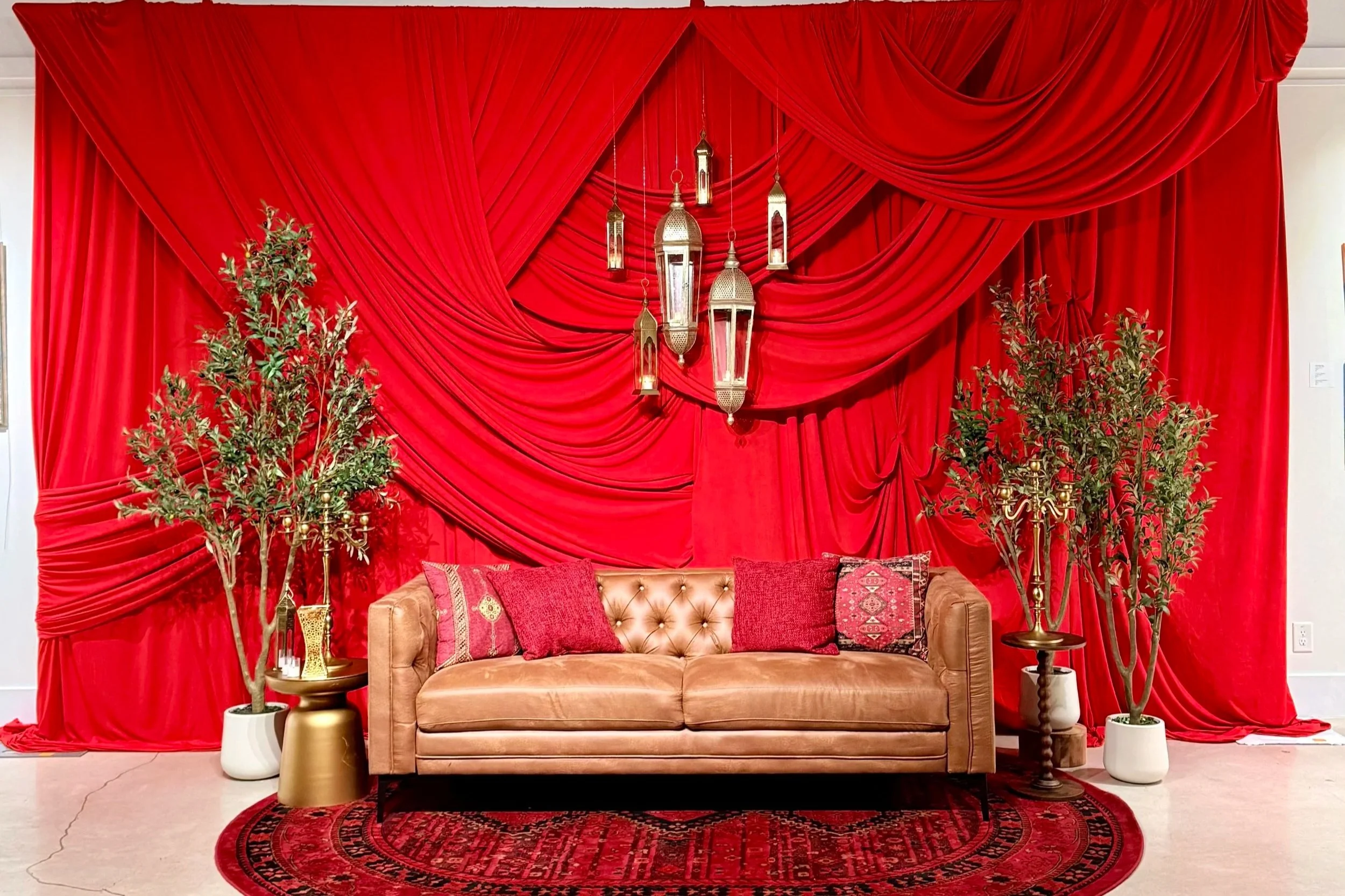 Empty art gallery room with paintings on the walls, a red curtain backdrop, a round red rug, a speaker, and white chairs in the foreground.