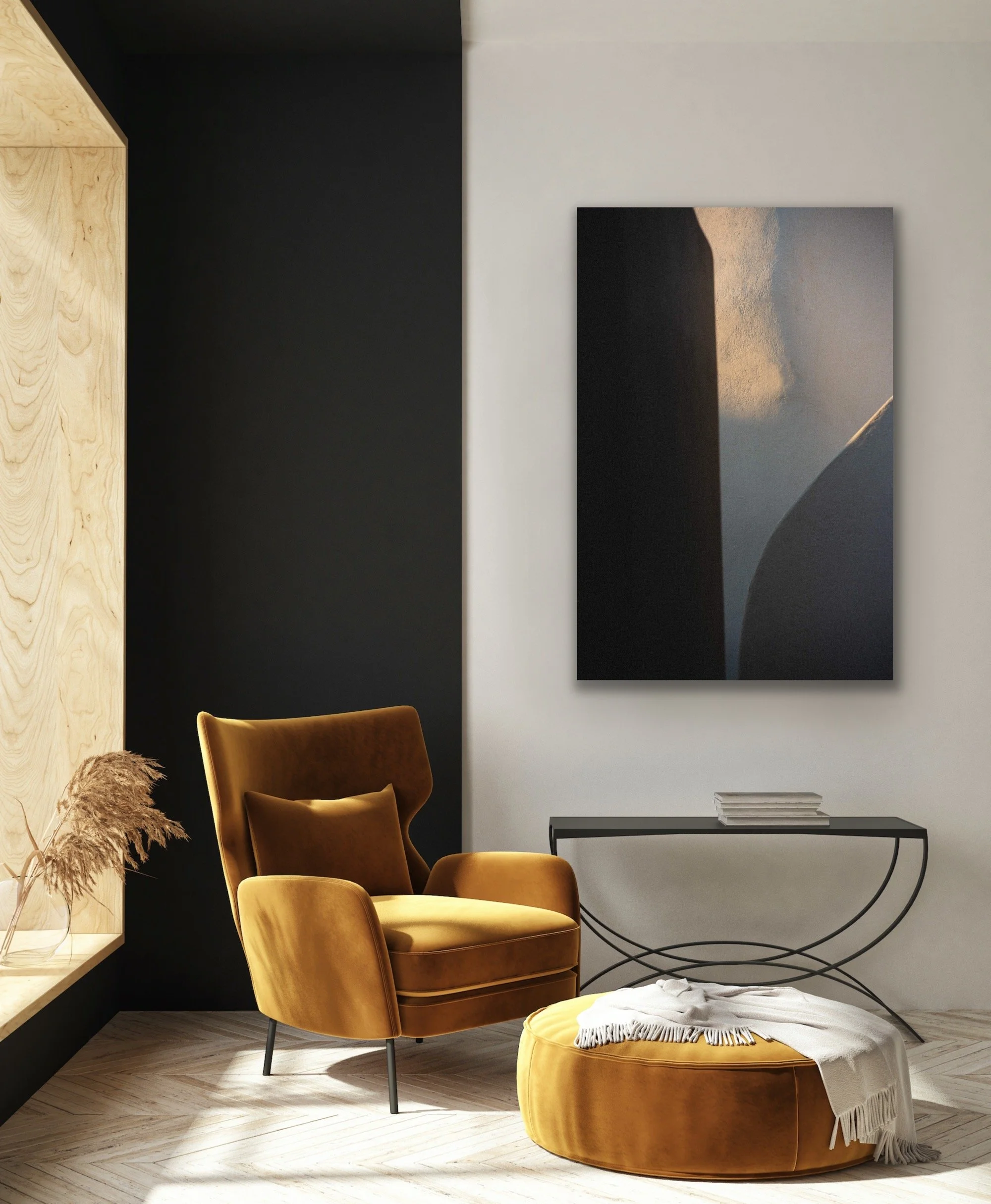 A cozy corner of a room with a vintage mustard-colored armchair and matching ottoman draped with a white throw blanket. There is a black side table with some stacked books, a large abstract painting on the white wall, and a window ledge with dried flowers against a black accent wall.