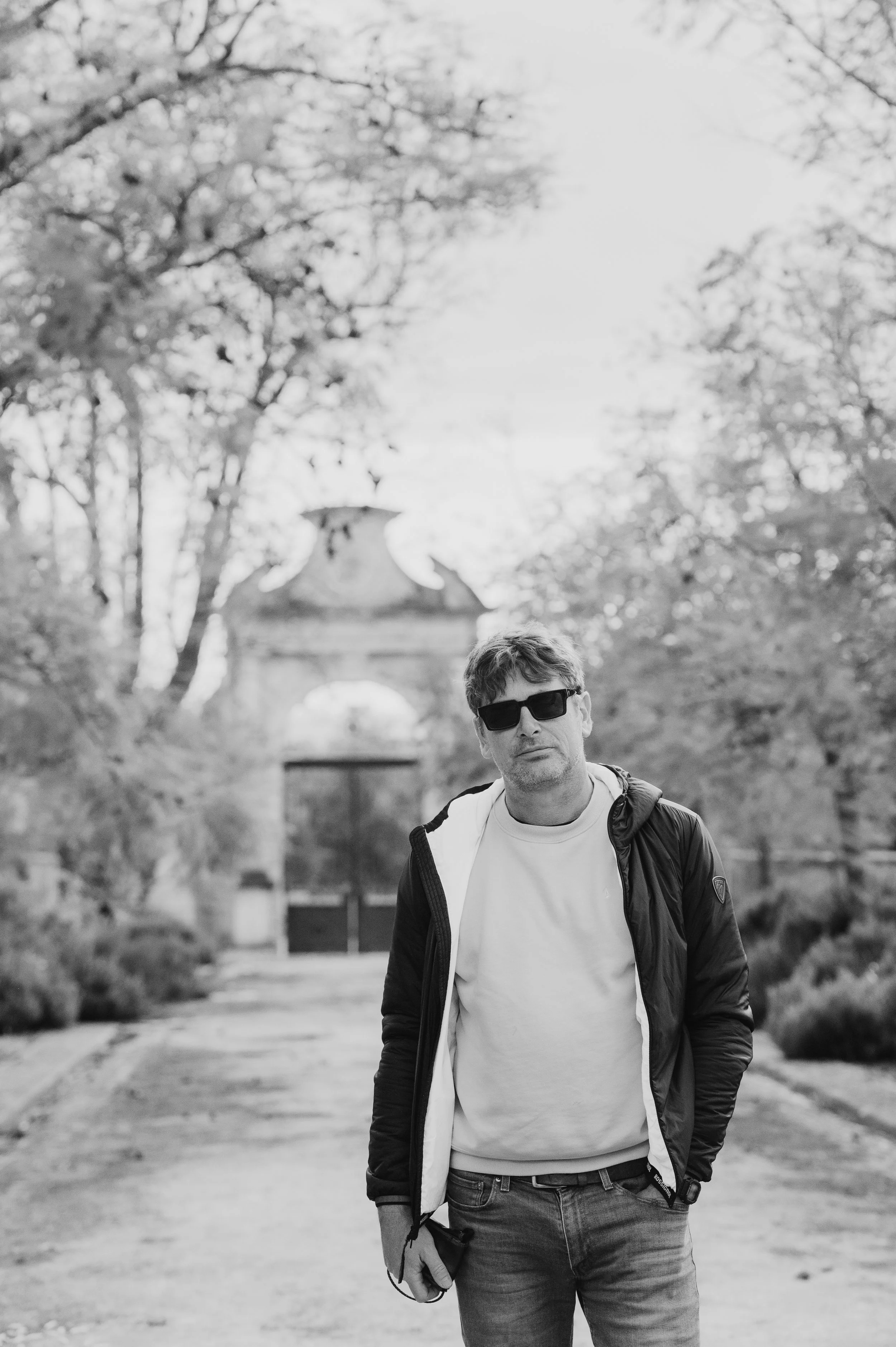 A man wearing sunglasses, a white t-shirt, a black jacket, and jeans, standing on a dirt pathway in a park with trees and a decorative archway in the background.