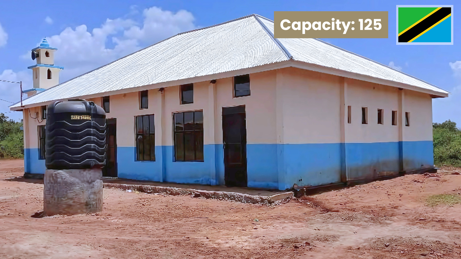 A light-colored building with a blue lower half and multiple windows, with a black water tank on a concrete stand outside, set on a dirt ground under a partly cloudy sky. The top right corner shows the Tanzanian flag, and text indicates a capacity of 125.