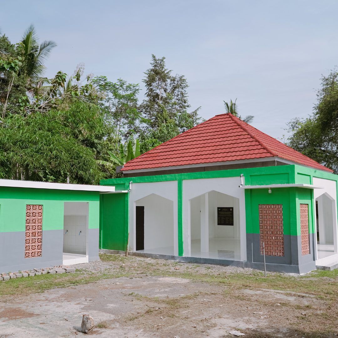 Small green and white building with a red tiled roof, surrounded by trees and a dirt ground.