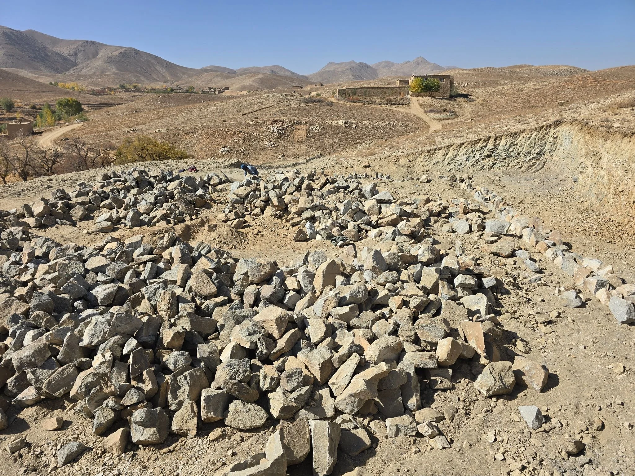 Archaeological excavation site with scattered rocks and dirt, surrounded by a dry, mountainous landscape under a clear blue sky.