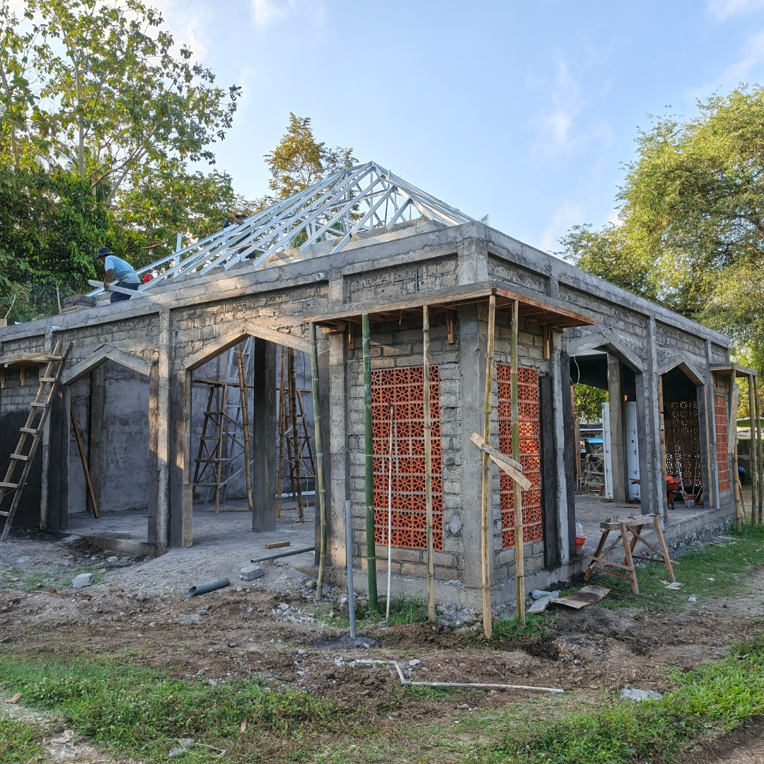 A house under construction with concrete and brick walls, workers on the roof installing trusses, and ladders leaning against the structure, surrounded by trees and construction tools.