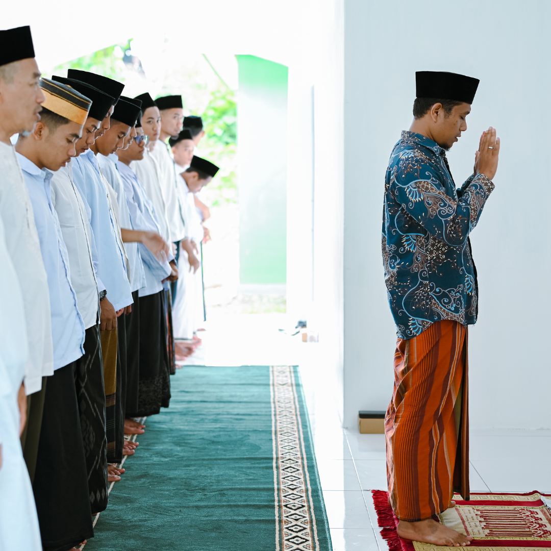 A group of men and boys praying together in a mosque or prayer room, with one man leading, all wearing traditional clothing and hats.